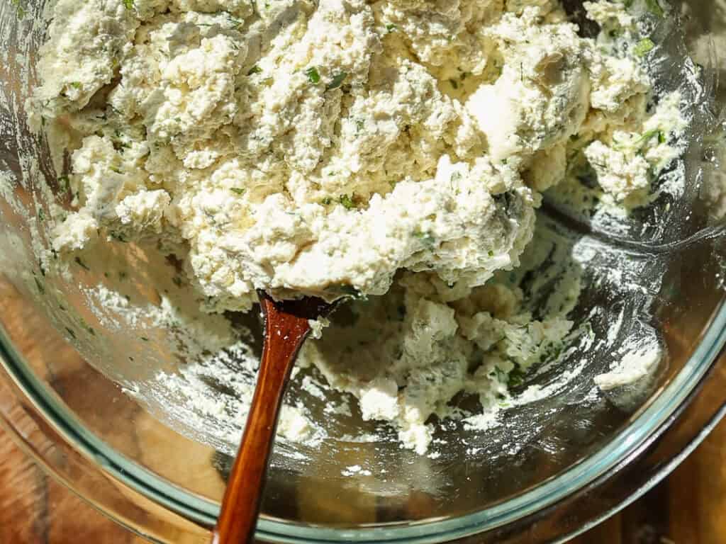 A glass bowl filled with fluffy ricotta cheese mixed with herbs. A wooden spoon is partially submerged in the mixture. The bowl is placed on a wooden surface, and bright sunlight highlights the texture and colors.